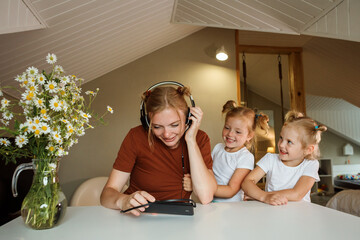 Mom is working, listening to music on her phone using earphones, her kids interfering her. - obrazy, fototapety, plakaty