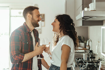 Young couple drinks a glass of white wine in the kitchen - couple in love celebrating their anniversary enjoying their new home - warm flare on background