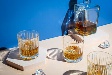 Whiskey with ice in glasses and bottle, beige and bright blue background with hard light, shadows...