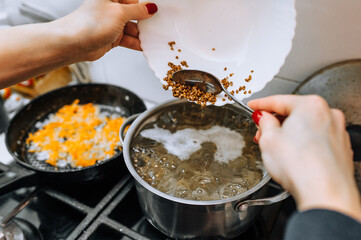 A woman, a cook, prepares soup in boiling water, pouring buckwheat from a plate into a saucepan. Cooking food in the kitchen.