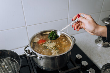 The girl, the cook, prepares soup from vegetables in boiling water, pouring green dill from a spoon into a saucepan.