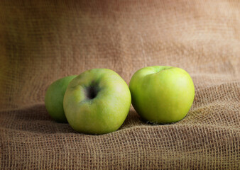 three green natural apples are on rough burlap under the light from the window