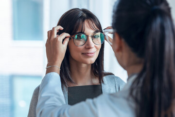 Female doctor choosing while proving eyeglasses to mature beautiful patient in medical consultation.