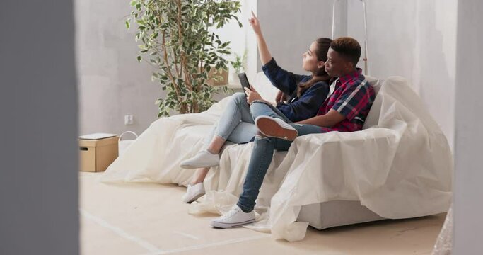 Couples of different nationalities are sitting on couch in living room, in newly purchased apartment during renovation, lovers discussing decoration furniture wall color watching projects on tablet