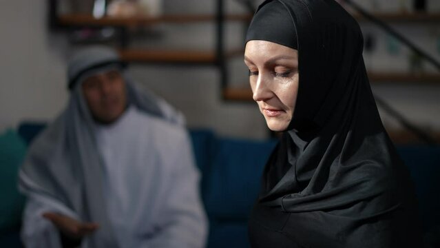 Side view portrait of stressed upset Middle Eastern woman in black hijab looking away thinking as man talking gesturing at background. Sad beautiful wife at home with husband arguing quarreling