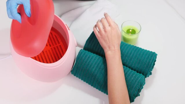 Close up video of a woman client hand lying on the towel with a glass of candle and cosmetologist opening paraffin box to make paraffin bath.