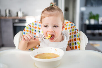 Cute baby girl toddler sitting in the high chair and eating her lunch soup at home kitchen.