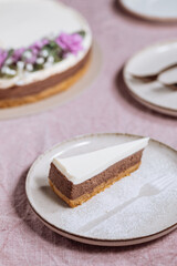 Chocolate cheesecake on bright plate with whole cake in blurry background. Decorated with flowers on pastel purple cloth. Vintage atmosphere, festive also. Juicy cake, one piece
