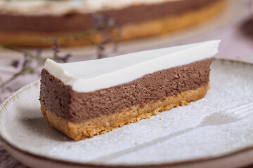 Chocolate cheesecake on bright plate with whole cake in blurry background. Decorated with flowers on pastel purple cloth. Vintage atmosphere, festive also. Juicy cake, one piece