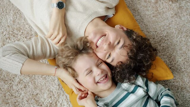 Top view of young lady and little boy laughing and looking at camera having fun lying on floor at home together. Emotion and happiness concept.