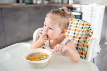 Cute baby girl toddler sitting in the high chair and eating her lunch soup at home kitchen.