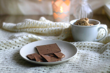 Plate with almond chocolate bar, mug with chocolate chip cookies, open book, reading glasses, soft blanket and lit candles. Hygge at home. Selective focus.