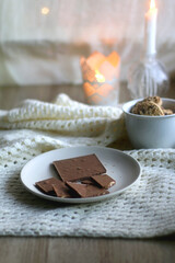Plate with almond chocolate bar, mug with chocolate chip cookies, open book, reading glasses, soft blanket and lit candles. Hygge at home. Selective focus.
