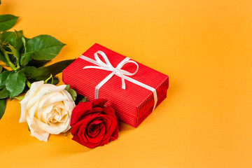 Red gift box with a white ribbon and two red and white roses over vibrant orange background. Valentine’s Day or Birthday gift concept