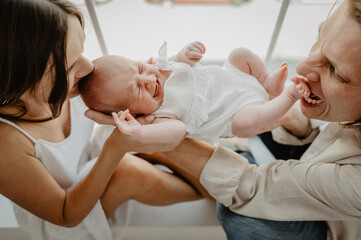 Obraz premium Young mom and dad are holding a newborn in their arms by the window. The family holds a newborn baby girl in their arms.