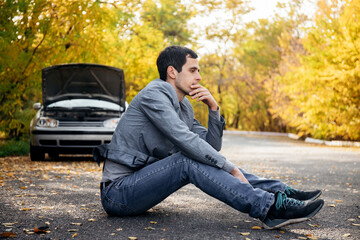 A man in sadness sits on the road in front of a broken car with an open hood