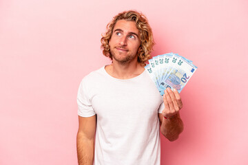Young caucasian man holding banknotes isolated on pink background dreaming of achieving goals and...