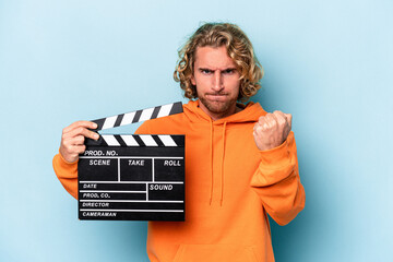 Young caucasian man holding a clapperboard isolated on blue background showing fist to camera,...
