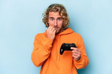 Fototapeta premium Young caucasian man playing with a video game controller isolated on blue background biting fingernails, nervous and very anxious.