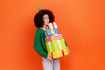 Amazed woman with Afro hairstyle wearing green casual style sweater standing with stack of presents in hands, being astonished to have lots gifts. Indoor studio shot isolated on orange background.