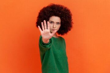 Fototapeta na wymiar Portrait of strict woman with Afro hairstyle wearing green casual style sweater showing stop ban gesture, looking at camera with serious expression. Indoor studio shot isolated on orange background.