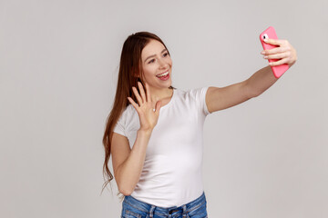 Portrait of happy friendly woman taking selfie and waving hand, gesturing hello, communicating on video call, online chatting, wearing white T-shirt. Indoor studio shot isolated on gray background.
