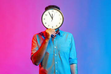 Portrait of man in shirt hiding face behind big wall clock display, wasting his time,...
