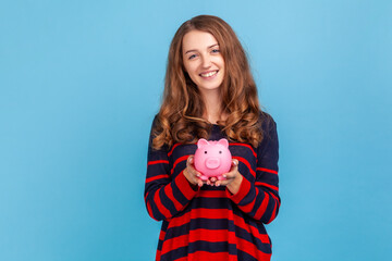 Fototapeta na wymiar Beautiful woman wearing striped casual style sweater, holding pink piggy bank, looking at camera with charming smile, best variant for saving money. Indoor studio shot isolated on blue background.