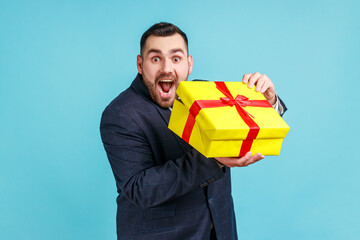 Excited man with beard wearing official style suit holding gift box, satisfied with holiday...