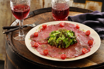 Prato de Carpaccio fatiado sobre barril de madeira rústica com copos de vinho tinto