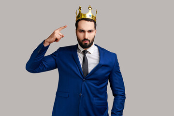 Self confident bearded businessman pointing fingers on golden crown on his head, showing his authority, wearing official style suit. Indoor studio shot isolated on gray background. - obrazy, fototapety, plakaty