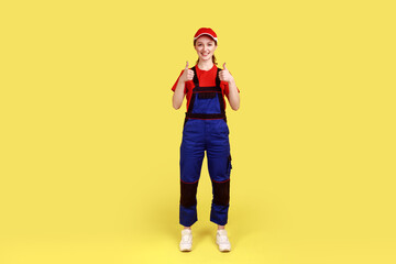 Ful llength portrait of worker woman standing and looking at camera, showing thumbs up, expressing happiness, wearing overalls and red cap. Indoor studio shot isolated on yellow background.