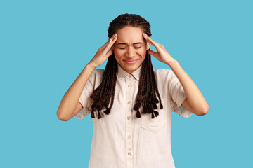 Unhappy distressed woman with black dreadlocks has high blood pressure, rubs temples to relieve headache, keeps eyes shut, has unbearable migraine. Indoor studio shot isolated on blue background.