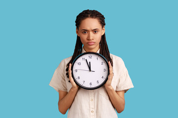 Portrait of serious woman with black dreadlocks holding big wall clock, looking at camera with...
