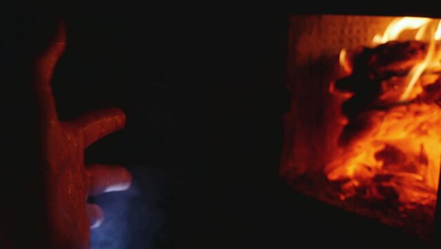 A man warms his hands after hypothermia while sitting by a warm hearth in the dark. Water vapor escapes from the skin. Extreme living conditions