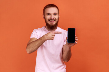 Portrait of bearded man pointing at cellphone and smiling at camera, recommending gadget or mobile application, wearing pink T-shirt. Indoor studio shot isolated on orange background.