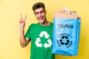 Young caucasian man holding a recycling bag full of paper to recycle isolated on yellow background joyful and carefree showing a peace symbol with fingers.