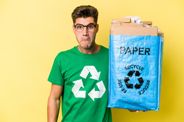 Young caucasian man holding a recycling bag full of paper to recycle isolated on yellow background shrugs shoulders and open eyes confused.