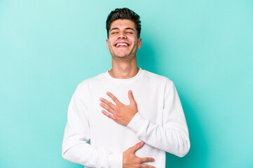 Young caucasian man isolated on blue background laughs happily and has fun keeping hands on stomach.