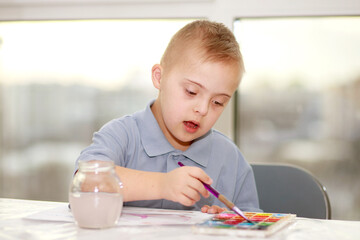 A boy with Down syndrome sits at a large table and draws with watercolors, the child develops and...