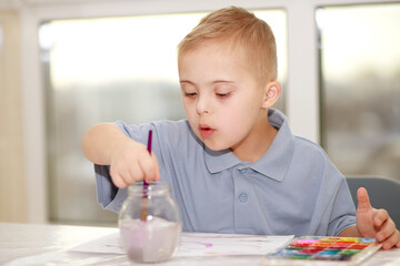 A boy with Down syndrome sits at a large table and draws with watercolors, the child develops and learns.
