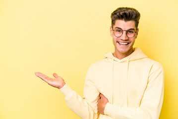 Young caucasian man isolated on yellow background showing a copy space on a palm and holding another hand on waist.