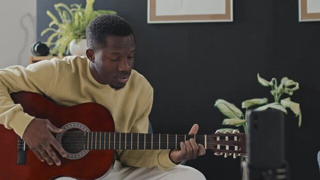 Medium Of Young Black Man Sitting In Armchair At Home At Daytime, Playing Acoustic Guitar, Smiling And Talking On Smartphone Camera, Streaming Live Video