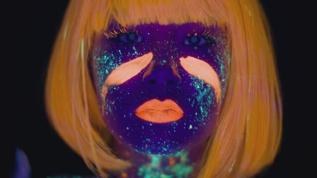 Neon body art. Close up of young woman wearing wig smearing orange paint on her cheeks, dancing in ultraviolet lights