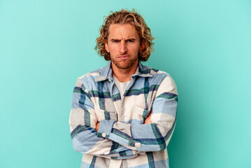 Young caucasian man isolated on blue background frowning face in displeasure, keeps arms folded.