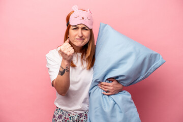 Young caucasian woman wearing a pajama holding pillow isolated on pink background showing fist to camera, aggressive facial expression.