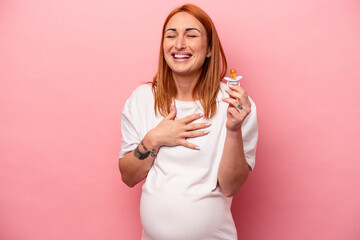 Young caucasian pregnant woman holding pacifier isolated on pink background laughs out loudly keeping hand on chest.