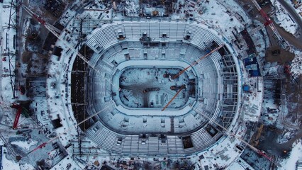 Snowy football field inside stadium with seats for spectators covered with snow and high steel building cranes pull out aerial view
