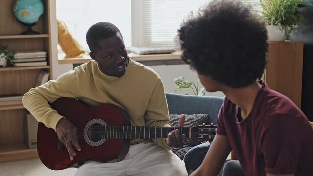 Side view of curly-haired young man sitting in armchair in living room at daytime, looking at Black male music teacher showing chords on acoustic guitar and talking