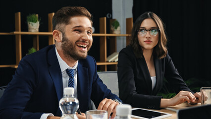 young businessman laughing near serious woman in eyeglasses during meeting